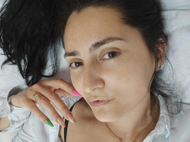 JennaEasy tem cabelo escuro, olhos castanhos e uma expressão ligeiramente séria. Ela está vestindo uma camisa branca, um colar dourado e tem unhas rosa, verdes e rosa, com um anel de ouro..