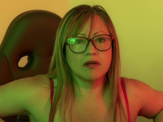 Christin Moore con iluminación verde y roja en su rostro, vistiendo una camiseta sin mangas roja, gafas y cabello rubio, luciendo sorprendida con los brazos levantados. Se ven los senos, de tamaño medio..