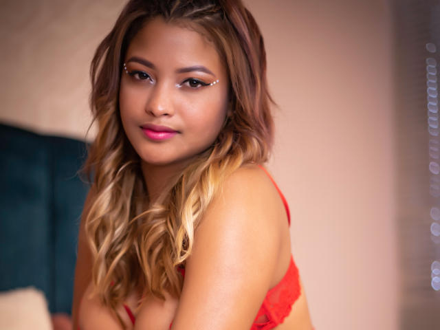 Kristal Jhonson, com cabelos loiros ondulados, lingerie vermelha e batom rosa, olhando para a câmera com um leve sorriso. Seu pequeno piercing no nariz e a maquiagem discreta são visíveis..