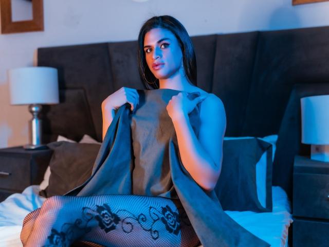 MissBritanny, con cabello oscuro, iluminación azul, vestida con un vestido gris, sosteniéndolo abierto para mostrar medias de encaje negro tipo "pescados", sentada en una cama, sosteniendo un cojín..