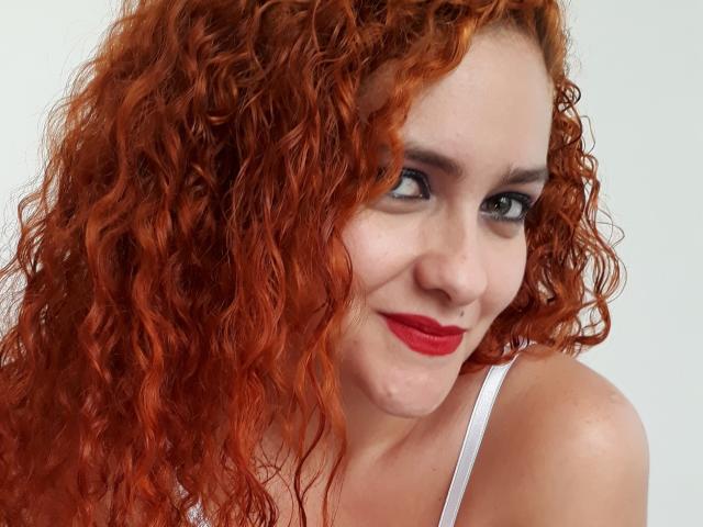 GinebraNova, con cabello rojo rizado, piel pálida, ojos verdes y lápiz labial rojo, sonriendo a la cámara, vestida con una blusa blanca, mostrando sus pechos y glúteos en la foto..
