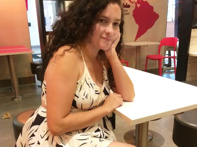 GinebraNova with curly brown hair, white and black patterned dress, sitting at a white table, resting her head on her hand, smiling slightly, in a diner with red chairs.