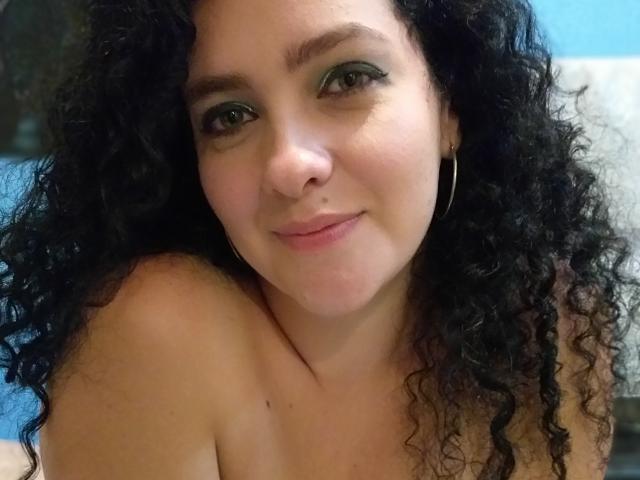 GinebraNova has curly black hair, green eye makeup, a nose ring, and is smiling with small, pink-tinted nipples visible. She's wearing a thin gold hoop earring.