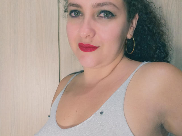 GinebraNova, con i capelli neri ricci, rossetto rosso, occhi verdi, orecchini a cerchio dorati e una maglia grigia scintillante che mostra il seno, con un leggero sorriso, su uno sfondo di legno..
