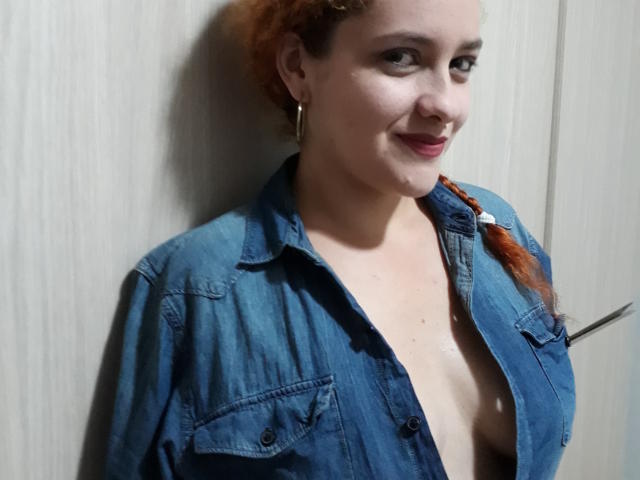 GinebraNova, con i capelli rossi intrecciati, una camicia di jeans blu e orecchini a cerchio dorati, sorride alla fotocamera, mostrando il seno, con uno sfondo di un muro di legno..