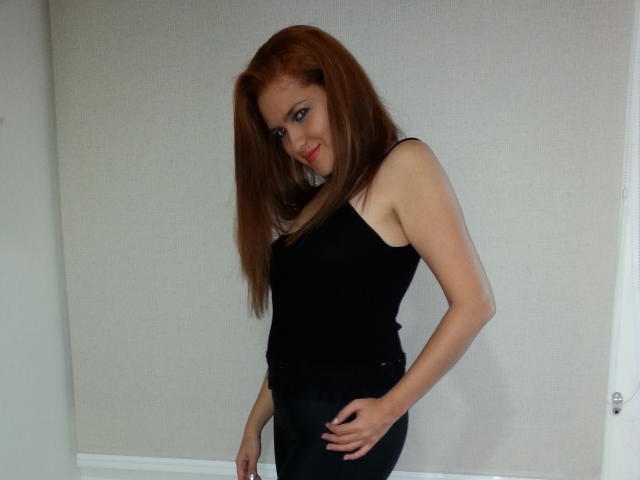 GinebraNova with red hair, black tank top, and black pants, smiling, standing, hand on hip, looking sideways, showing small tits, fair skin, and a cute face.
