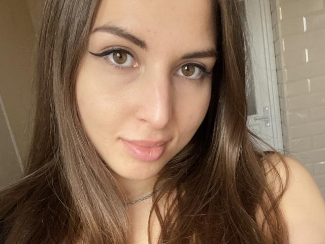 DominikaRos a les yeux marron, de longs cheveux bruns, et elle fait une moue en retroussant légèrement ses lèvres roses. Elle porte un eye-liner noir en forme d'aile et un collier fin..
