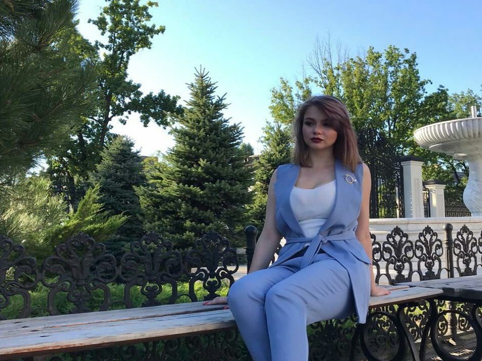SofiaLeena, vêtue d'une robe bleu clair, d'un haut blanc et d'une ceinture assortie, assise sur un banc en bois avec une structure en métal noir ornée, montrant ses seins, dans un jardin ensoleillé..