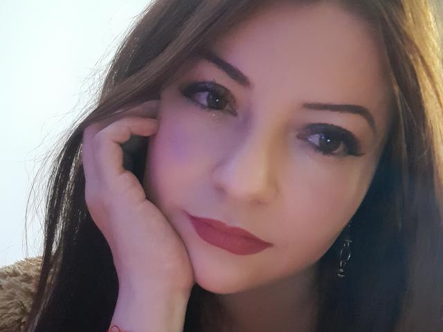 NatalyHami with red lipstick, brown eyes, and dark brown hair, resting her hand on her cheek, showing a slight smile, wearing a furry brown garment.