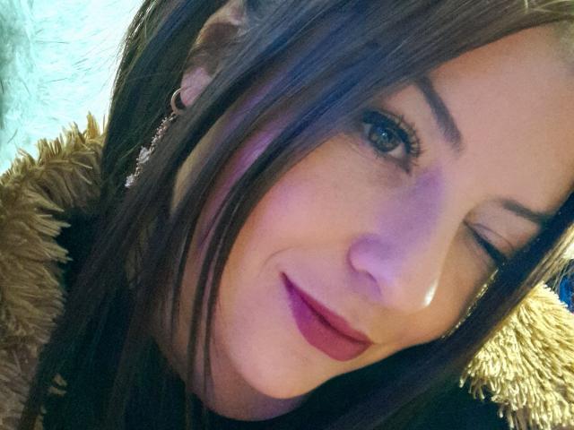 NatalyHami with winking brown eyes, long brown hair, purple lipstick, and a cute smile, wearing a brown furry jacket. Her tits are not visible. She has a cute face.