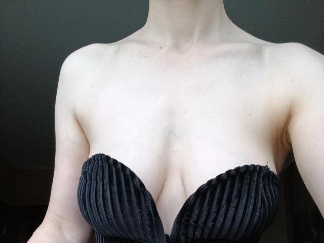 PenJanis, con piel pálida, lleva un vestido negro sin tirantes con textura, que muestra sus pechos, con un ligero escote, clavícula visible y parte superior del pecho. Sus brazos se ven parcialmente a los lados..