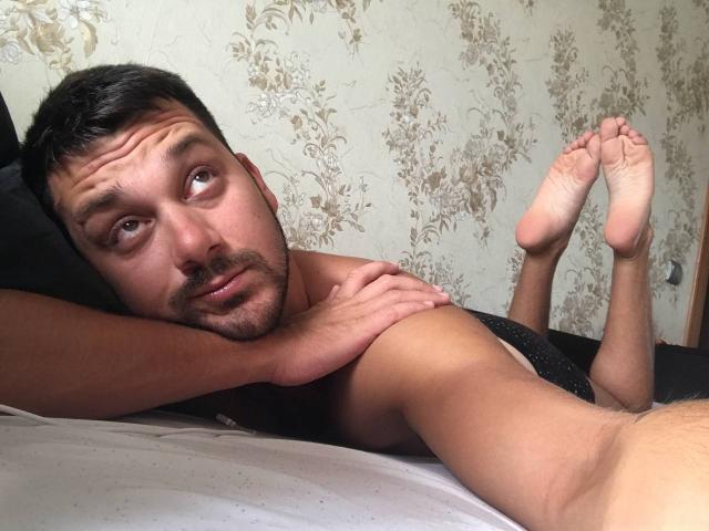 Maeltra liegt ohne Hemd auf einem Bett, die Beine angezogen, die Fußsohlen sind sichtbar, die Arme sind über dem Kopf verschränkt, er hat kurzes, dunkles Haar und einen leicht verwirrten Gesichtsausdruck. Seine Hand ruht auf seinem Arm..