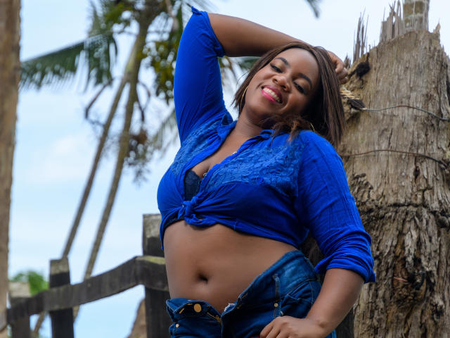 DanielaBrown with dark skin, blue tied-up shirt showing her tits, black bra, unbuttoned jeans, palm tree background, arm raised, smiling, red lipstick, sexy, confident.