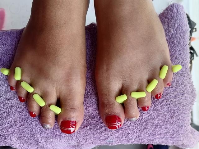 De voeten van VioletaSwitch, met felrode teennagels en neongele gel-teensteun, rusten op een paarse handdoek. Haar huid is donkerbruin..