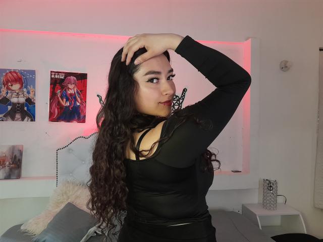 Antonnella Tyler, avec de longs cheveux noirs bouclés, portant un haut noir à manches longues, touchant sa tête d'une main, montrant ses fesses et ses seins, dans une chambre avec des posters d'anime..