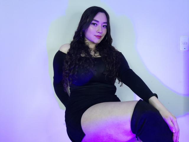 AntonnelaTyler with long dark hair, black off-shoulder dress, and thigh-high boots, sitting with a slight smile, purple and blue lighting on her thighs.