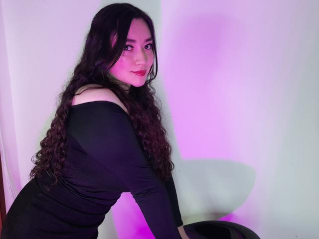 Antonnella Tyler, com longos cabelos cacheados e pretos, vestindo uma blusa preta com um ombro de fora, inclinada para frente, olhando por cima do ombro, com iluminação rosa e roxa no rosto e uma parede branca..
