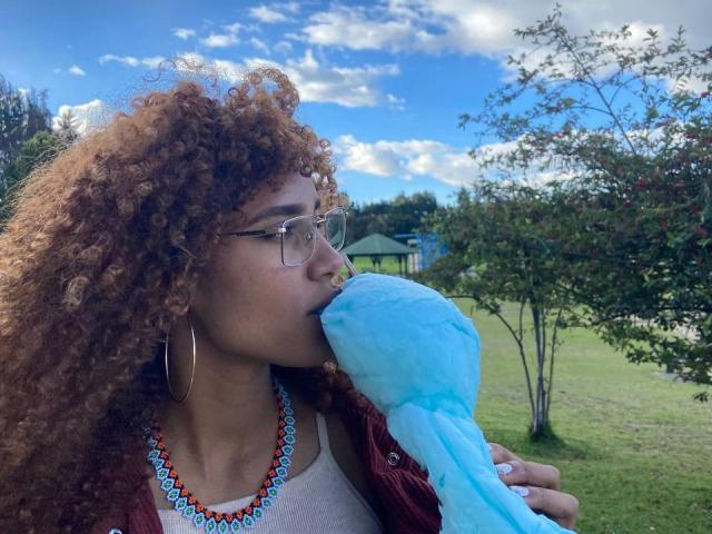 CinthiaWalker with curly brown hair, glasses, nose ring, hoop earrings, and colorful necklace, sucking on a blue fluffy toy, standing in a grassy outdoor area with trees.
