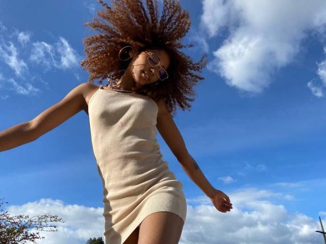 Cinthia Walker, con i capelli castano ricci, indossa un vestito beige senza maniche, ha un seno piccolo e porta gli occhiali. È in piedi di fronte a un cielo blu con nuvole bianche, con le braccia aperte e lo sguardo rivolto verso il basso..