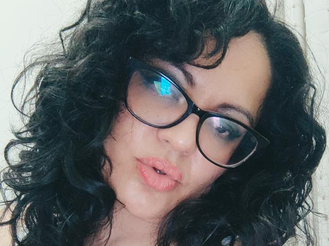 Mya Collins tem cabelos pretos encaracolados, olhos azuis grandes e está fazendo beicinho com os lábios rosados levemente entreabertos. Ela está usando óculos pretos e olhando para a câmera..