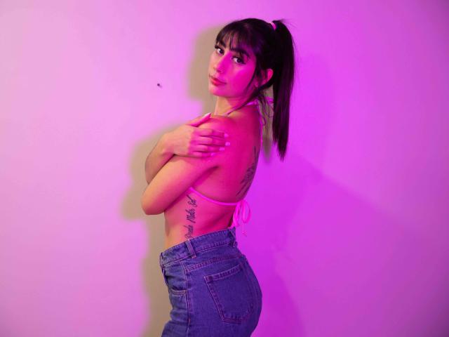 JessicaSoler met een paardenstaart, armen gekruist, roze bh, blauwe jeans, tatoeage op de rechter ribbenkast, paars-roze verlichting, kijkt over haar schouder, toont kleine borsten en billen..