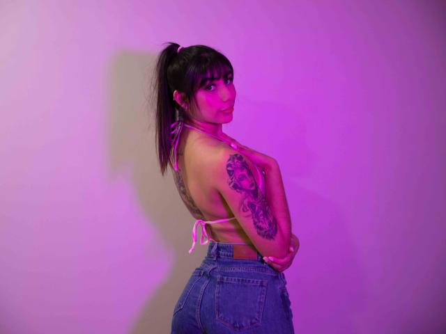 JessicaSoler with a ponytail, pink bikini top, and blue jeans, showing her back tattoo, looking over her shoulder, pink and purple lighting, highlighting her ass and tits.