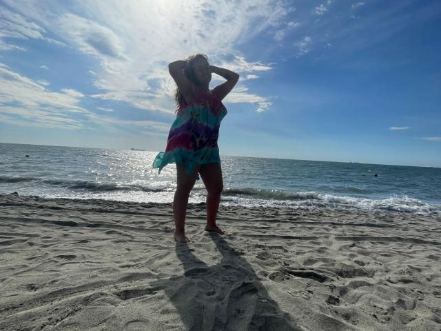 Joanallen met armen omhoog, gekleed in een kleurrijke tie-dye jurk, staat op een zandstrand, de zon schijnt, de lucht is blauw en er is een oceaan op de achtergrond, er is een schaduw op het zand..