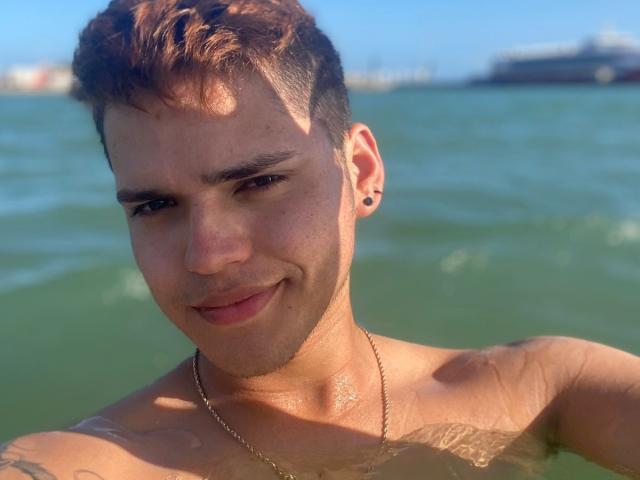 EddTerence, senza camicia, con capelli castani corti, piercing alle orecchie e una catena d'argento, sorride in acqua verde, con la luce del sole sulla sua pelle bagnata, e ha dei tatuaggi sulla parte superiore del braccio..