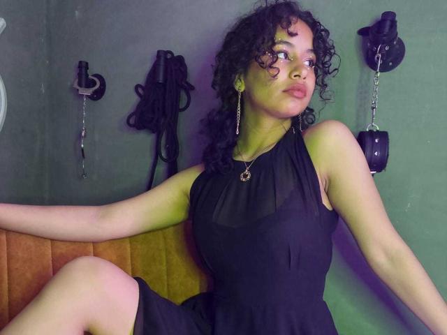 Heily Peterson, con cabello rizado, vestido negro y collar de cadena, sentada con un brazo extendido, mostrando sus senos, en un sofá naranja, mirando hacia arriba, con juguetes sexuales negros en el fondo..