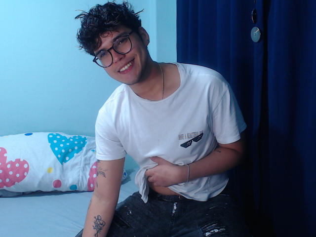 EddTerence with curly black hair, glasses, white t-shirt, and torn jeans, smiling, holding shirt, tattoo on left arm, standing by bed with colorful pillow.