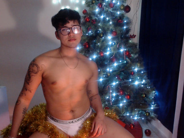 EddTerence, ohne Hemd, mit Brille, in weißen Boxershorts, mit goldener Deko, mit Tattoos, sitzt vor einem Weihnachtsbaum mit blauen Lichtern und roten Ornamenten. EddTerence hat ein süßes Gesicht..
