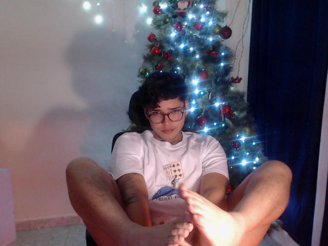 EddTerence mit lockigem, schwarzem Haar, Brille, weißem T-Shirt und Tattoo, sitzt mit hochgezogenen Beinen, die Füße zeigen zur Kamera. Im Hintergrund steht ein Weihnachtsbaum mit Lichtern und Dekorationen..