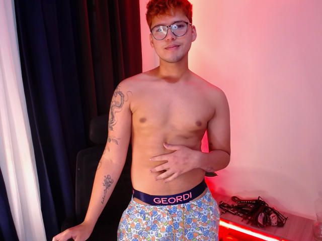 EddTerence ohne Hemd, mit Brille, kurzen roten Haaren und Tätowierungen am linken Arm, trägt blaue Boxershorts mit einem "GEORDI"-Bündchen, steht mit einer Hand auf dem Bauch und hat ein leichtes Lächeln..