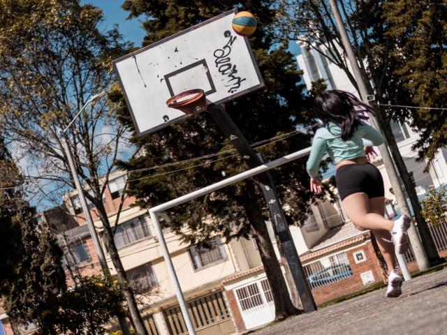 MelanyEtThomi, met zwart haar en een groene top, springt om een basketbal in het net te gooien, gekleed in zwarte shorts en witte sneakers, op een buitenbaan met een verweerde basketring..