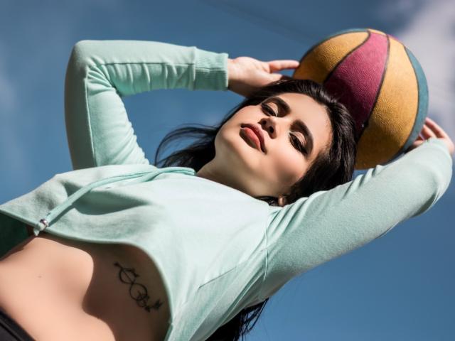 MelanyEtThomi mit dunklem Haar, die einen mintgrünen Reißverschluss-Pullover trägt, hält einen bunten Ball über ihrem Kopf und zeigt dabei ein Tattoo auf ihrem Unterbauch, vor einem strahlend blauen Himmel..