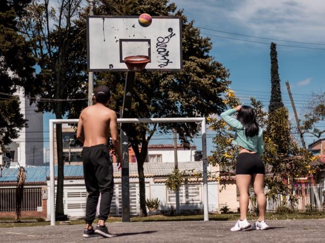 MelanyEtThomi, ohne Hemd, schwarzer Trainingsanzug, Ball in der Hand, mit dem Rücken zur Kamera, stehend neben dem Basketballkorb. MelanyEtThomi, grünes Oberteil, schwarze Shorts, weiße Turnschuhe, wirft den Ball. Draußen, tagsüber..