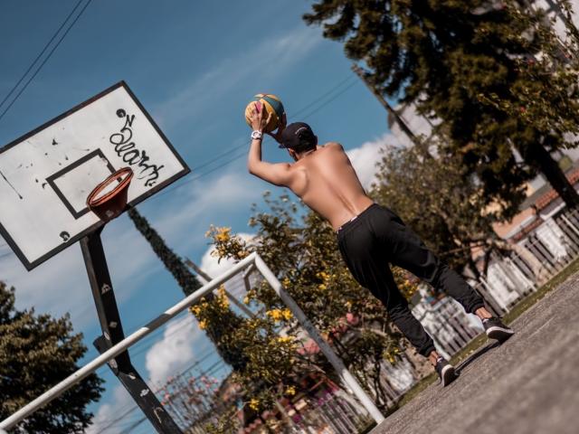 MelanyEtThomi, torse nu, portant un pantalon noir et une casquette, tient un ballon de basket au-dessus de sa tête, dos à la caméra, sur un terrain de basket extérieur ensoleillé avec un panier..