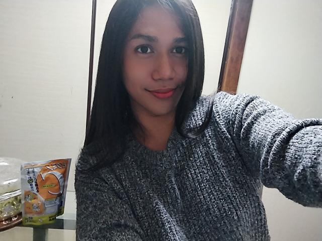 ShinjukuTS with medium brown skin and long black hair, wearing a gray knit sweater, smiling with red lips, holding a phone for a selfie. Pack of oranges on left.