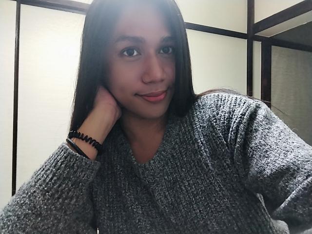 ShinjukuTS con cabello largo y oscuro, vistiendo un suéter gris de punto, sonriendo ligeramente, con la cabeza apoyada en la mano, una pulsera negra en la muñeca, luciendo adorable..