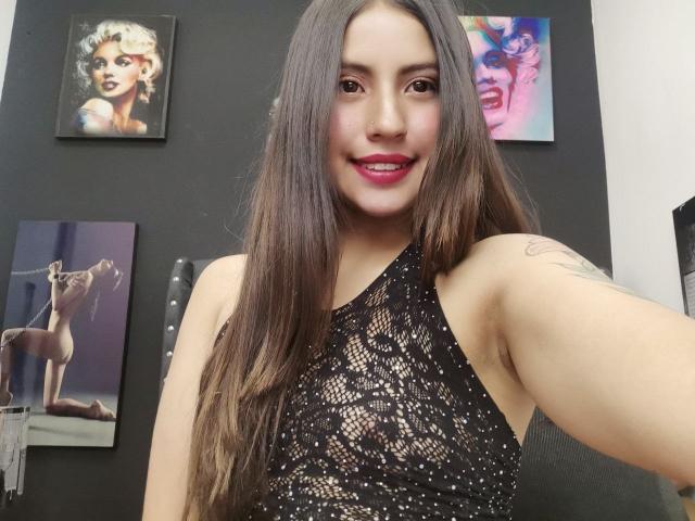 PaulaCeleste with long brown hair, black lace top, showing her tits, smiling, pink lipstick, tattoo on right arm, three paintings in background.