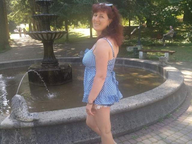 BellisimaAh mit roten Locken, einem blauen, gepunkteten Kleid, steht neben einem Steinfountain, zeigt ihr Gesäß und lächelt, hält eine Sonnenbrille auf dem Kopf, in einem Park..