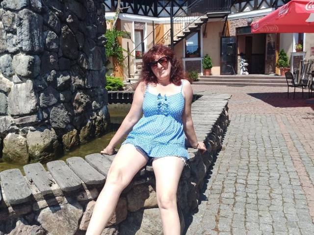 BellisimaAh with curly red hair, wearing a blue polka dot dress, sits on a stone bench, legs spread, showing her pale thighs, with her small tits and smiling face.