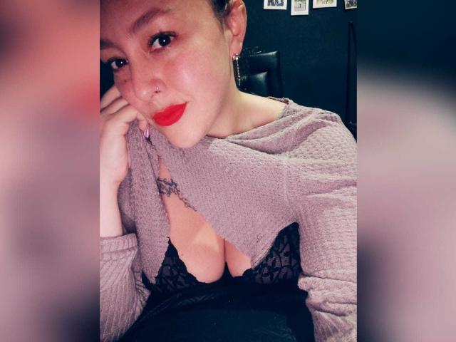 Bordette, con lápiz labial rojo, lleva puesto un top de punto morado, dejando ver un sujetador de encaje negro y sus pechos. Tiene una sonrisa encantadora, aretes y está apoyando la cabeza en su mano..
