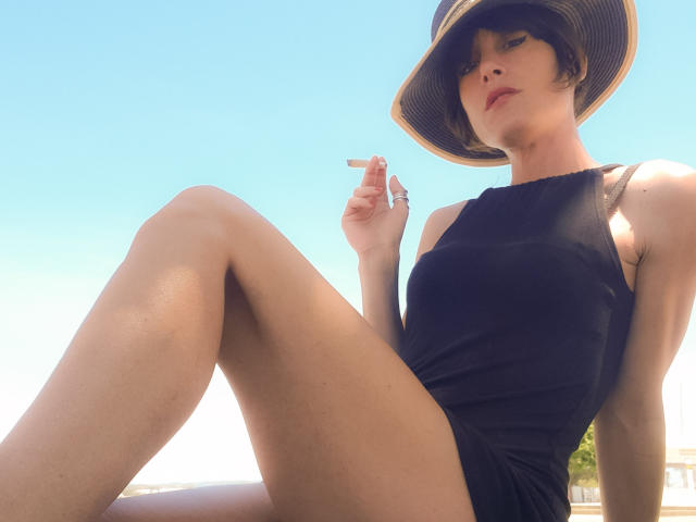 Aiyana with short brown hair, wearing a black sleeveless dress and straw hat, sits with legs crossed, holding a cigarette, showing her ass and smooth skin under a clear blue sky.