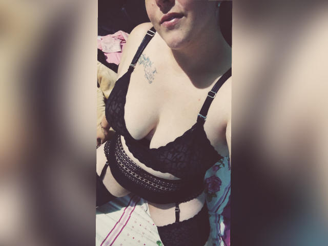 Bordette con un sujetador de encaje negro, una faja y medias, mostrando sus pechos y un pequeño tatuaje en el lado derecho de su pecho, de pie con una ligera sonrisa. Primer plano borroso..