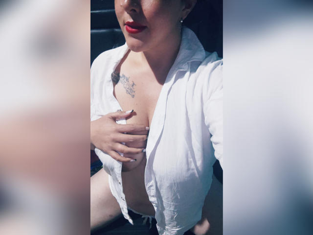 Vrouw met rode lipstick, open wit shirt, hand die haar borsten bedekt, tatoeage op de borst, zwarte nagels, zittend in een auto, zonlicht op haar gezicht, licht onscherpe randen..