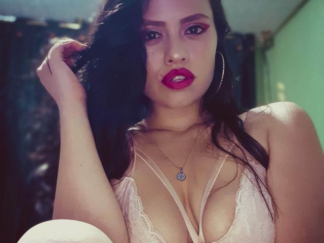 BellaSmithX69 with dark hair, pink lipstick, and hoop earrings, wearing a pink lace bra that shows her big tits, holding her hair, and a necklace with a pendant.