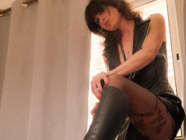 Aiyana with shoulder-length brown hair, wearing a black sleeveless top, black thigh-high boots, and sheer black stockings with floral patterns, sitting with knees up, looking down.