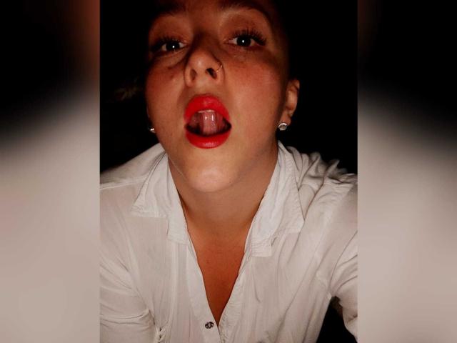 Femme avec rouge à lèvres rouge et la langue tirée, chemise blanche, boucles d'oreilles de perles, bouche légèrement ouverte, piercing au nez, fond sombre, regardant vers le haut, lumière sur le visage, cheveux bruns courts..