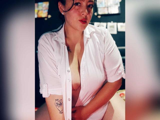 Bordette vestindo uma camisa branca, aberta para mostrar os seios, segurando a camisa para baixo, com uma tatuagem no braço, lábios rosados, cabelo escuro, brincos, em uma foto ligeiramente borrada..